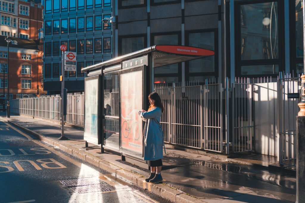 photographie d'une passante qui attend à un arrêt de bus, on peut y voir divers panneaux de signalisation et des publicités affichés sur l'abribus